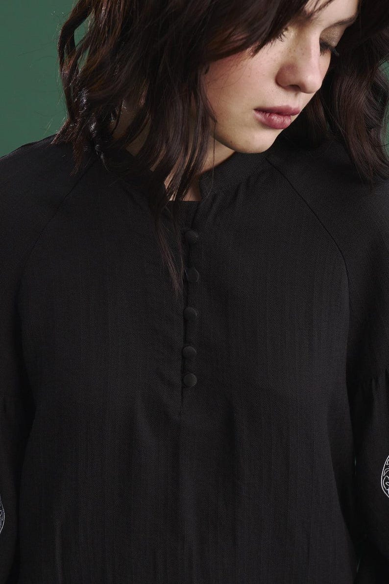 May include: Close-up of a black blouse with a mandarin collar and a series of four buttons down the front. The sleeves are long and slightly flared. The fabric has a subtle texture. The blouse is a solid black color.