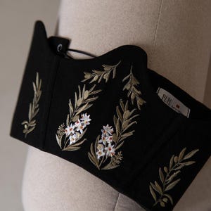 May include: A black corset top featuring intricate embroidery of olive branches and small white flowers with orange centres. The garment has a scalloped neckline and a white interior label with the text PUPU printed in black lettering.