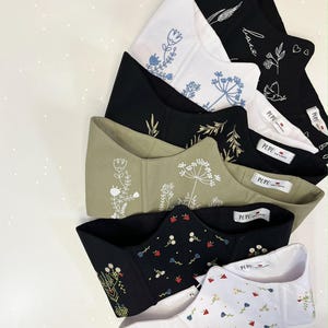 May include: A collection of six corset-style tops featuring various embroidered floral and botanical designs. The garments come in black, white, and olive green, each with a white interior label reading PUPU. The embroidery includes white, blue, and multi-colored thread patterns.