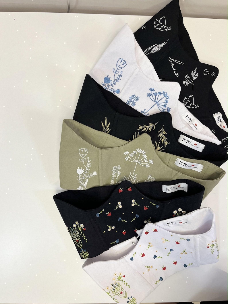 May include: A collection of six corset-style tops featuring various embroidered floral and botanical designs. The garments come in black, white, and olive green, each with a white interior label reading PUPU. The embroidery includes white, blue, and multi-colored thread patterns.