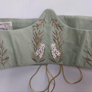 May include: A sage green corset top featuring embroidered olive branches and small white flowers. The garment has a pointed front hem, structured seams, and a white interior label with the text Hrushevka. Thin gold-toned laces hang from the bottom.