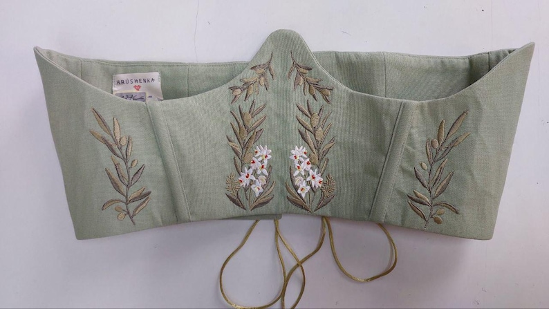 May include: A sage green corset top featuring embroidered olive branches and small white flowers. The garment has a pointed front hem, structured seams, and a white interior label with the text Hrushevka. Thin gold-toned laces hang from the bottom.