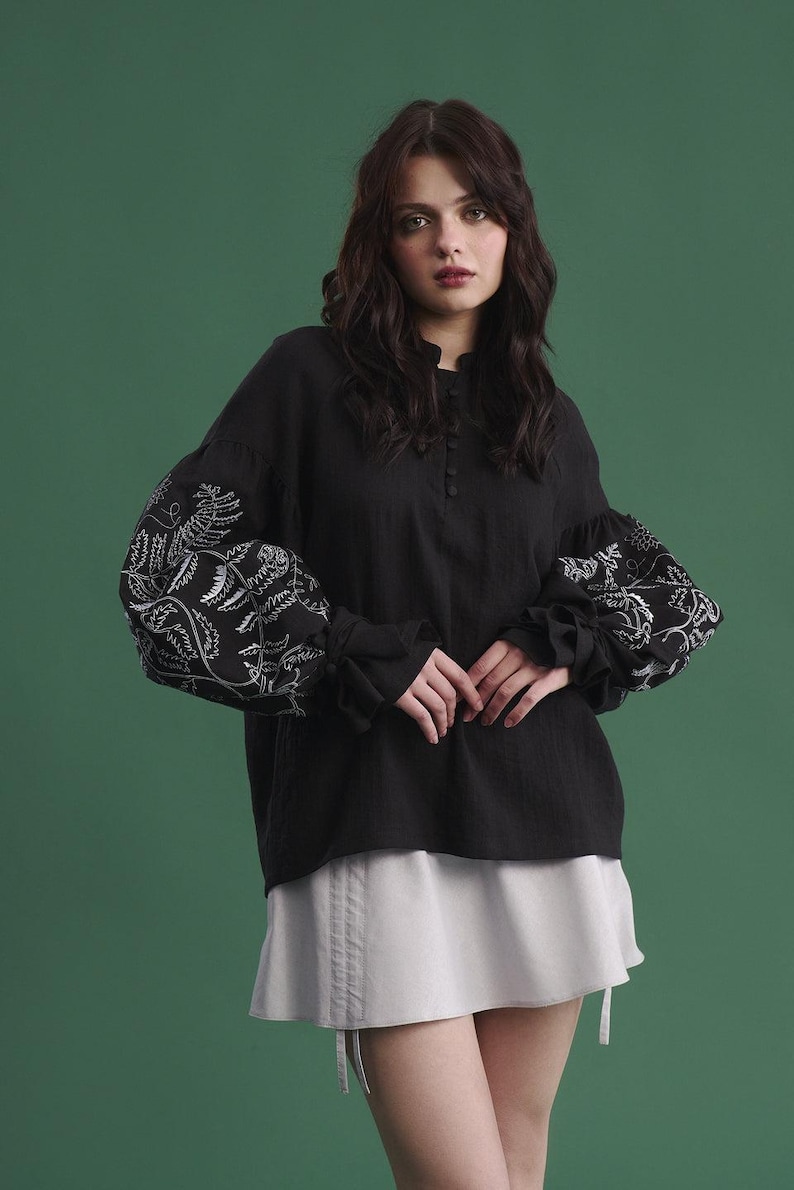 May include: Black long-sleeve blouse with white floral embroidery on the sleeves. The blouse has a high neckline and button details. Paired with a light gray mini skirt. The background is a solid green.