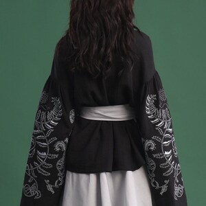 May include: Black top with white floral embroidery on the sleeves, paired with a white skirt and a white belt. The sleeves are long and billowy. The background is a solid green.