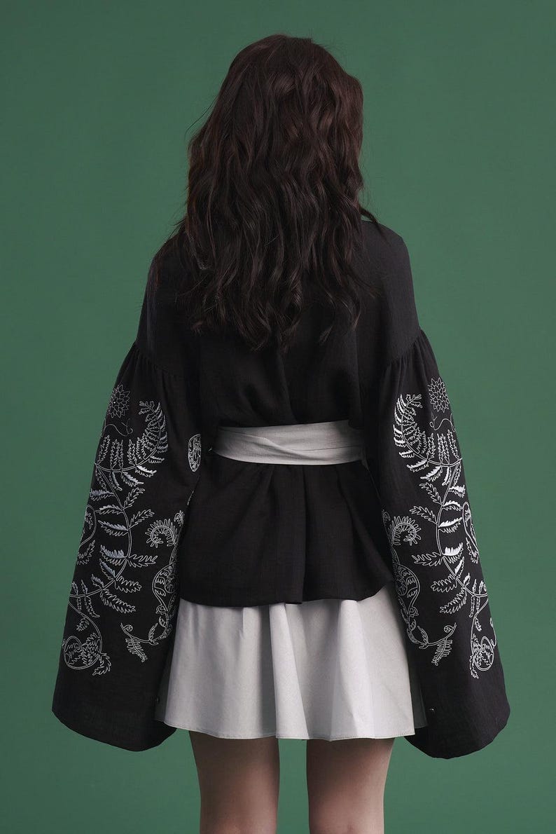 May include: Black top with white floral embroidery on the sleeves, paired with a white skirt and a white belt. The sleeves are long and billowy. The background is a solid green.