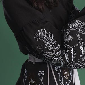 May include: Black long-sleeved top and skirt with white embroidered fern and floral designs. The top features a buttoned front and flared sleeves. The skirt is a light gray color. The design includes a black apron-like piece with embroidered heart and fern motifs.