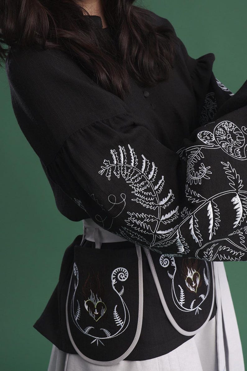 May include: Black long-sleeved top and skirt with white embroidered fern and floral designs. The top features a buttoned front and flared sleeves. The skirt is a light gray color. The design includes a black apron-like piece with embroidered heart and fern motifs.