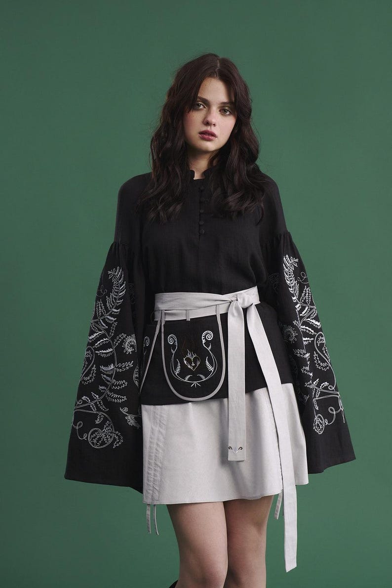 May include: Black top with embroidered sleeves and a matching skirt. The sleeves feature white floral embroidery. A gray apron-style belt with a decorative design and a matching tie cinches the waist. The outfit is set against a green backdrop.