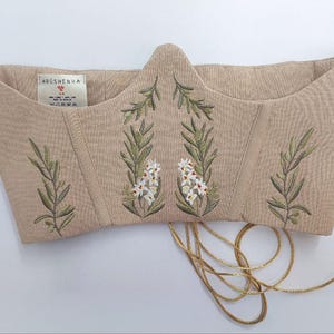 May include: A beige corset top featuring embroidered green olive branches and white flowers. The garment has a pointed front neckline, vertical structural seams, and thin beige laces hanging from the bottom. A white label reads HRUSHENKA.