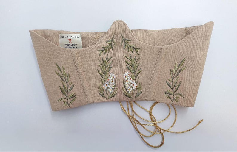 May include: A beige corset top featuring embroidered green olive branches and white flowers. The garment has a pointed front neckline, vertical structural seams, and thin beige laces hanging from the bottom. A white label reads HRUSHENKA.