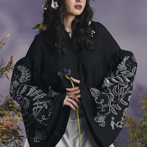 May include: Black blouse with embroidered white fern and floral designs on the sleeves. The blouse has a button-down front and wide sleeves. The model is wearing a white skirt and has flowers in their hair.