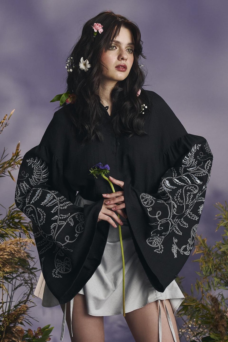 May include: Black blouse with embroidered white fern and floral designs on the sleeves. The blouse has a button-down front and wide sleeves. The model is wearing a white skirt and has flowers in their hair.