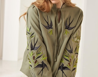 May include: An olive green long-sleeve blouse featuring a high neckline with a tie closure and elasticized cuffs. The garment is decorated with detailed embroidery of dark grey swallows, bright green leaves, and small brown olives on the sleeves.