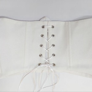 May include: A white strapless corset top featuring a central vertical lace-up closure with silver-toned metal eyelets and white ribbon lacing. The garment has structured vertical seams and a straight hemline, with loose ribbon ends hanging below.