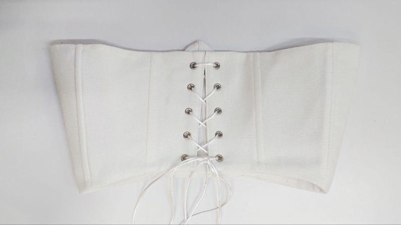 May include: A white strapless corset top featuring a central vertical lace-up closure with silver-toned metal eyelets and white ribbon lacing. The garment has structured vertical seams and a straight hemline, with loose ribbon ends hanging below.