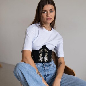 May include: A white short-sleeved t-shirt worn over a black corset featuring gold and white floral embroidery. The corset has a structured, curved hemline and is paired with light blue denim jeans. The outfit combines casual and structured elements.