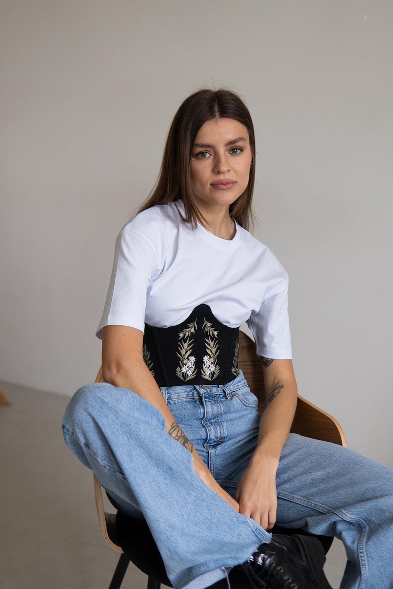 May include: A white short-sleeved t-shirt worn over a black corset featuring gold and white floral embroidery. The corset has a structured, curved hemline and is paired with light blue denim jeans. The outfit combines casual and structured elements.