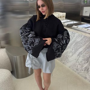 May include: A black blouse with white floral embroidery on the sleeves and a light blue skirt. The blouse has a high neckline and button details. The skirt has drawstring details. The person is wearing sunglasses and yellow heels.