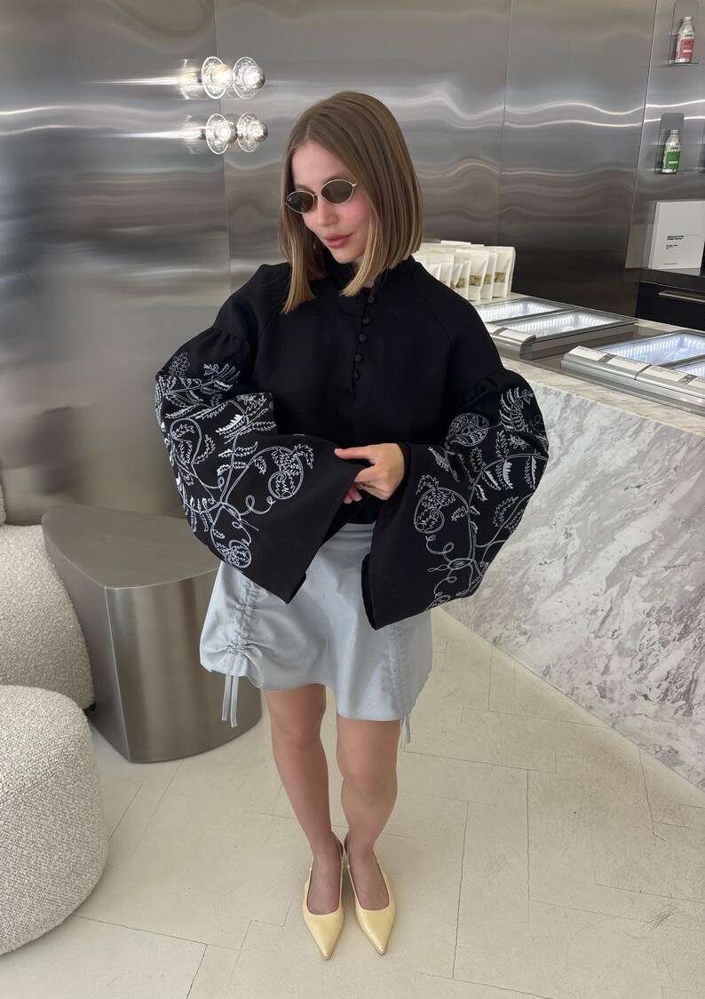 May include: A black blouse with white floral embroidery on the sleeves and a light blue skirt. The blouse has a high neckline and button details. The skirt has drawstring details. The person is wearing sunglasses and yellow heels.