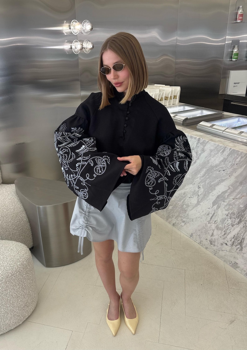 May include: A black blouse with white floral embroidery on the sleeves and a light blue skirt. The blouse has a high neckline and button details. The skirt has drawstring details. The person is wearing sunglasses and yellow heels.