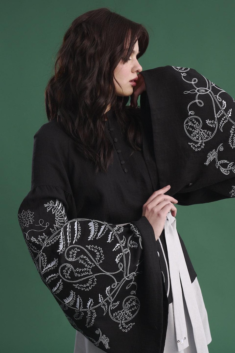 May include: Black long-sleeved top with white floral embroidery on the sleeves. The top has a high collar and button details. A white sash is tied around the waist. The background is a solid green.