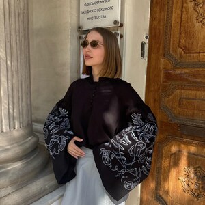 May include: A black blouse with wide sleeves featuring white floral embroidery. The blouse is paired with a light gray skirt. The image is taken outdoors in front of a wooden door.