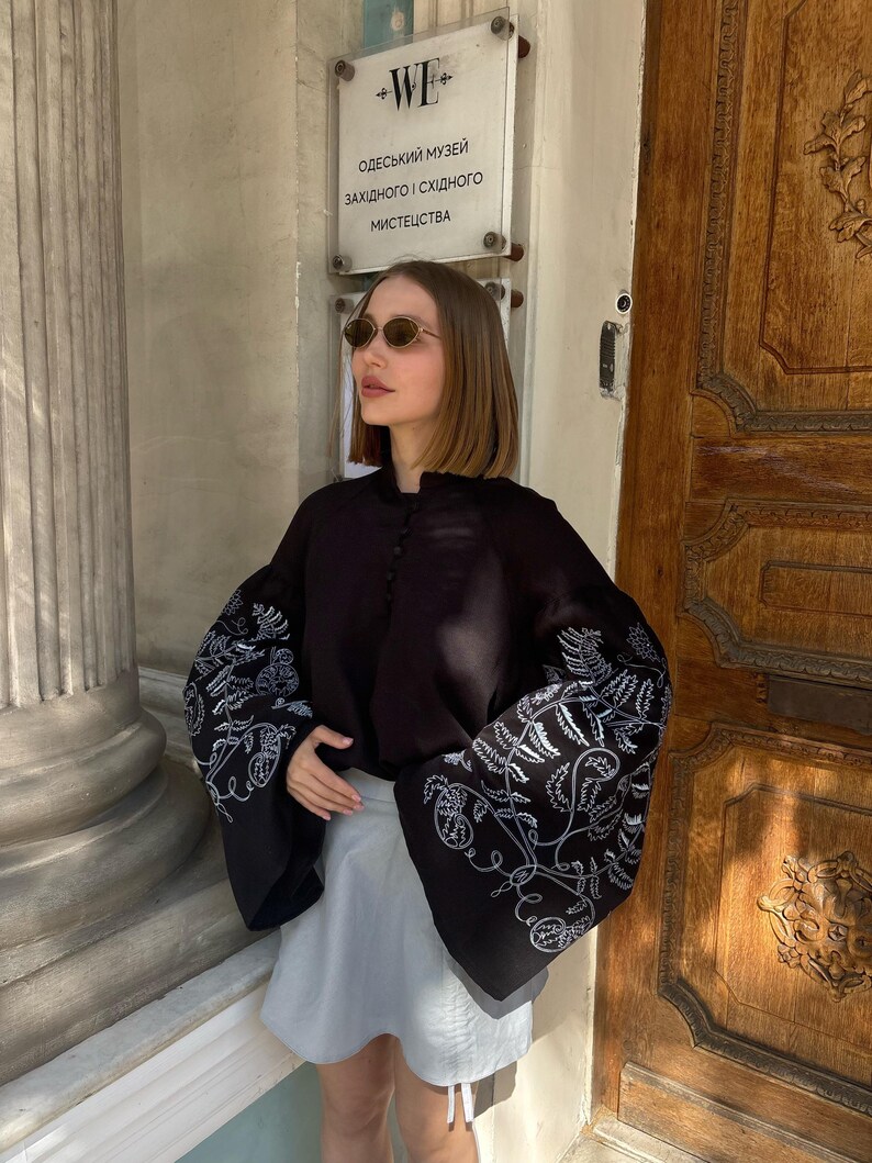 May include: A black blouse with wide sleeves featuring white floral embroidery. The blouse is paired with a light gray skirt. The image is taken outdoors in front of a wooden door.