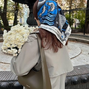 May include: A square headscarf featuring a vibrant blue, black, and white graphic print of stylized birds and geometric patterns, worn over hair. The scarf is paired with a solid olive green shoulder bag and a beige trench coat.
