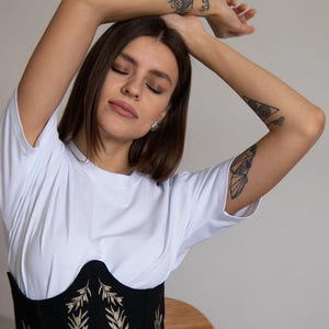 May include: A white short-sleeved t-shirt worn under a black corset-style bodice featuring embroidered floral patterns in shades of green, white, and gold, paired with light blue denim jeans, creating a layered outfit with a structured waist.