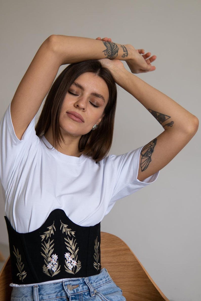 May include: A white short-sleeved t-shirt worn under a black corset-style bodice featuring embroidered floral patterns in shades of green, white, and gold, paired with light blue denim jeans, creating a layered outfit with a structured waist.