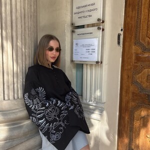 May include: A black blouse with white floral embroidery on the sleeves. The blouse has a high collar and button details. The image also shows a sign with Ukrainian text, including museum hours.