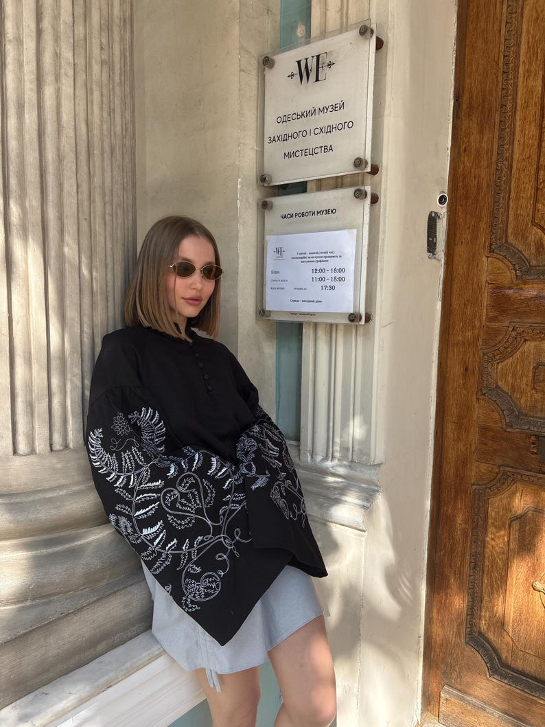 May include: A black blouse with white floral embroidery on the sleeves. The blouse has a high collar and button details. The image also shows a sign with Ukrainian text, including museum hours.