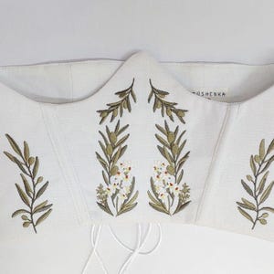 May include: A white corset top featuring embroidered olive branches with green leaves and small white flowers. The garment has a structured, pointed neckline and includes thin white laces hanging from the bottom edge. The brand name OSHENKA is visible.