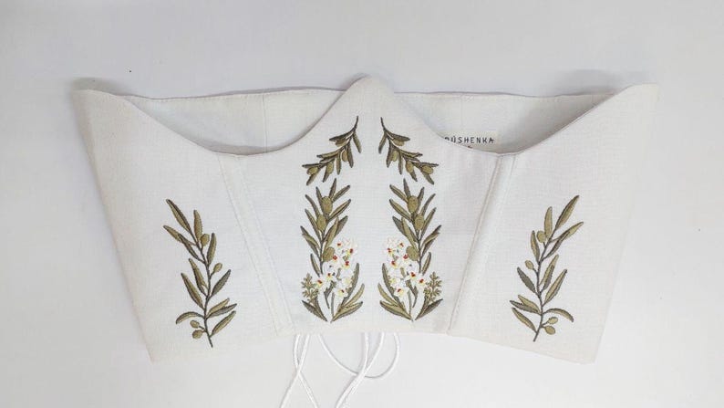 May include: A white corset top featuring embroidered olive branches with green leaves and small white flowers. The garment has a structured, pointed neckline and includes thin white laces hanging from the bottom edge. The brand name OSHENKA is visible.
