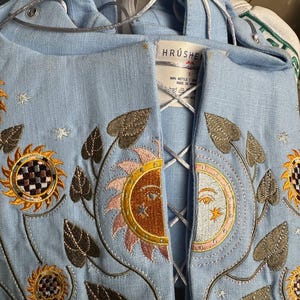 Peut inclure: Haut en lin bleu clair avec broderies de soleil, lune et tournesols. Le haut présente un devant lacé et des coutures décoratives. L'étiquette de la marque "HRUSHE" est visible. La broderie comprend des accents dorés, marron et roses.