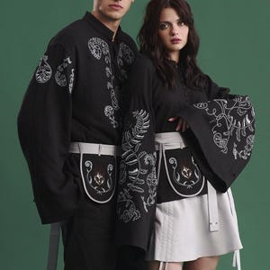 May include: Two people wearing matching black outfits with white embroidered floral designs. The outfits include long-sleeved shirts and skirts or pants, with matching white aprons. The background is a solid green.