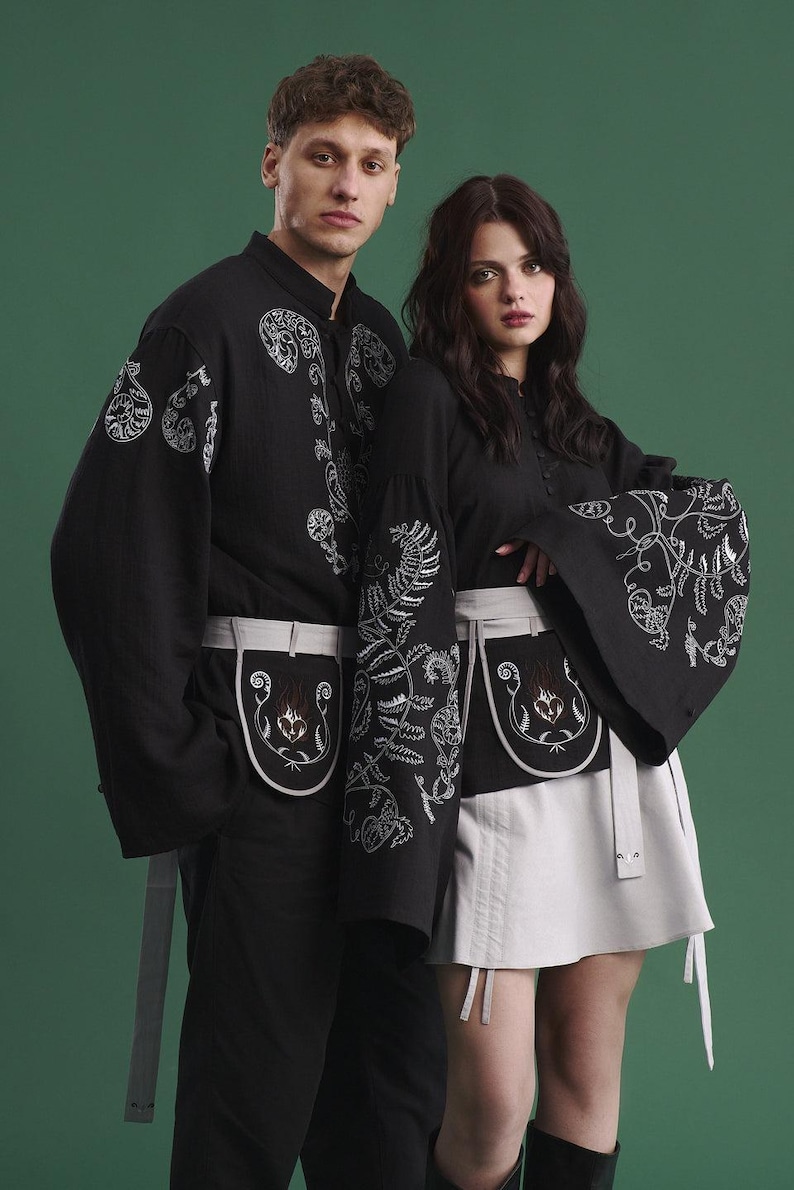 May include: Two people wearing matching black outfits with white embroidered floral designs. The outfits include long-sleeved shirts and skirts or pants, with matching white aprons. The background is a solid green.