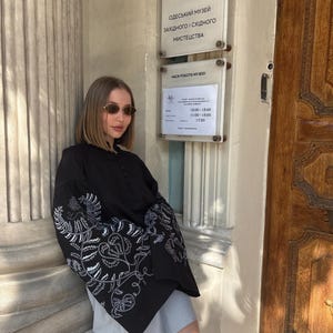 May include: A black blouse with white floral embroidery on the sleeves. The blouse is paired with a gray skirt. The image also shows a museum sign with Ukrainian text.