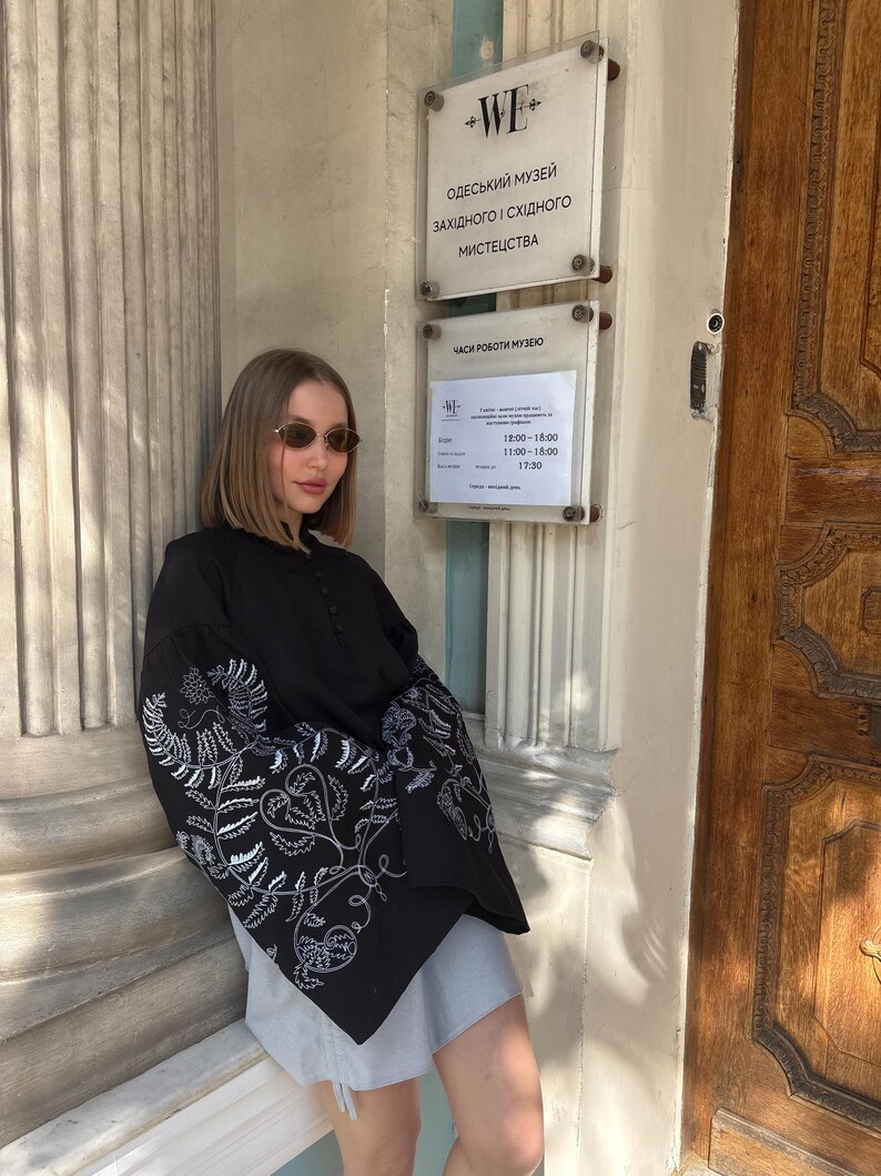 May include: A black blouse with white floral embroidery on the sleeves. The blouse is paired with a gray skirt. The image also shows a museum sign with Ukrainian text.