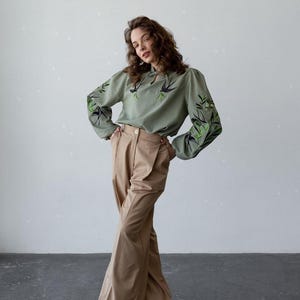 May include: A sage green long-sleeve blouse featuring a keyhole neckline and dark bird and leaf embroidery on the chest and sleeves, paired with high-waisted tan wide-leg trousers that have a front button closure and side pockets.
