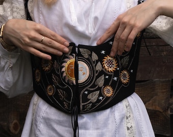 May include: A black corset featuring intricate embroidery of a sun and moon face at the center, surrounded by smaller suns, stars, and leaf motifs. The garment has a front lace-up closure with thin black cords and wide shoulder straps.