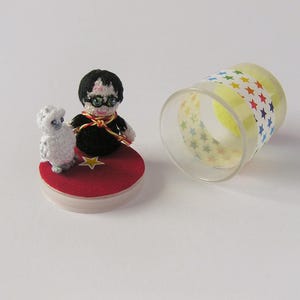 May include: A handmade crochet figurine of a character with glasses and a black outfit, and a white sheep, on a red base with a gold star. A clear plastic cup with a star pattern is next to the figurines.