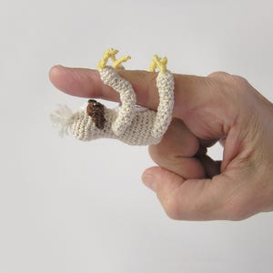 May include: A beige crocheted sloth finger puppet with yellow claws and a brown patch, hanging from a finger. The puppet's handmade details are visible. The design is simple and charming, ideal for imaginative play and storytelling.