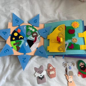 May include: A felt quiet book featuring a circular spinner with numbered blue triangles from two to seven, a sun, and various icons. The open page displays a yellow door, a green tree, and a sun. Loose felt finger puppets include a wolf, a cow, a koala, and a bearded figure.