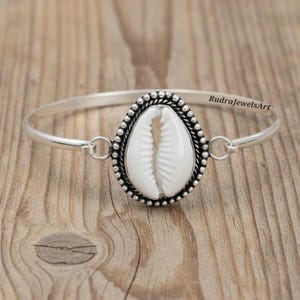 May include: A silver bangle bracelet with a white seashell centrepiece. The shell is set in an oval frame with a beaded border. The bracelet has a simple, elegant design.