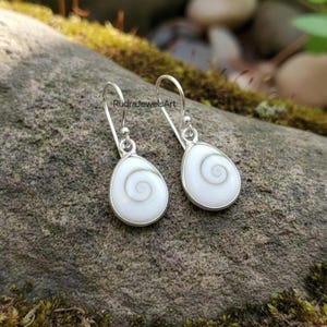 May include: A pair of silver drop earrings with a teardrop-shaped white pendant. Each pendant features a spiral design in the center. The earrings are displayed on a mossy rock. The text "RudraJewelsArt" is visible.