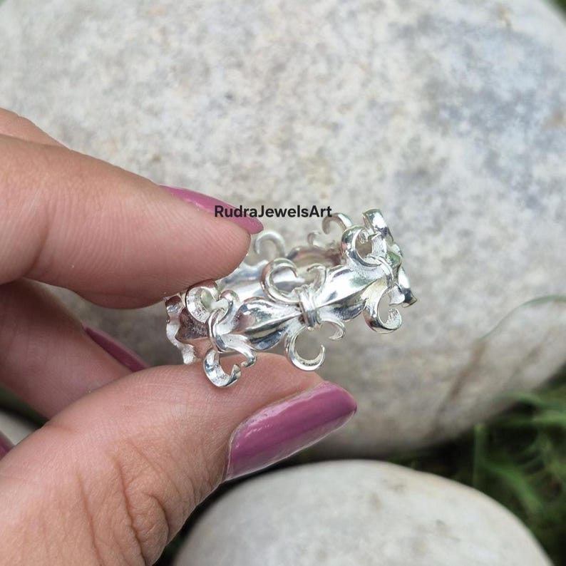 May include: A silver ring with an ornate, floral design. The ring features a repeating pattern of stylized flowers and curves. The ring is held in a hand with painted nails. The text "RudraJewelsArt" is visible.