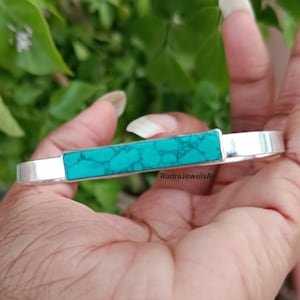 May include: A silver bracelet with a rectangular turquoise stone centerpiece. The turquoise stone has a mottled pattern. The bracelet is held in a hand against a blurred green background.