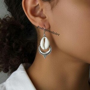 Puede incluir: Pendiente plateado con un colgante de concha blanca y una base en forma de media luna. El pendiente cuelga de un gancho y presenta un diseño detallado y punteado alrededor de la media luna. El texto "RudraJewelsArt" es visible.