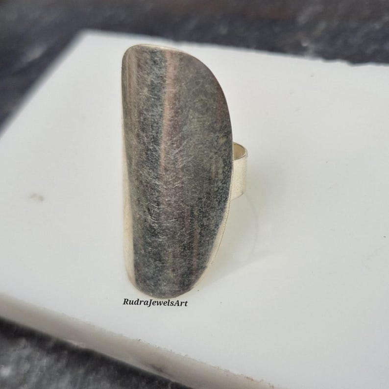 May include: A silver-toned ring with a large, elongated oval face. The face of the ring has a textured, striped appearance with shades of gray and silver. The ring is set on a white surface, and the brand name "RudraJewelsArt" is visible.